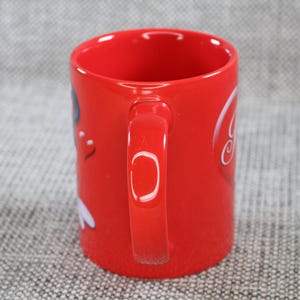 Peut inclure: Une tasse en c&eacute;ramique rouge avec une anse incurv&eacute;e. La tasse pr&eacute;sente un motif graphique blanc sur le c&ocirc;t&eacute;. La tasse est cylindrique et a une finition brillante. Le fond est une surface grise textur&eacute;e.