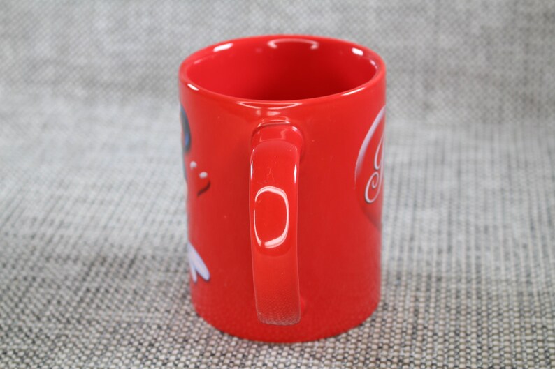 Peut inclure: Une tasse en c&eacute;ramique rouge avec une anse incurv&eacute;e. La tasse pr&eacute;sente un motif graphique blanc sur le c&ocirc;t&eacute;. La tasse est cylindrique et a une finition brillante. Le fond est une surface grise textur&eacute;e.
