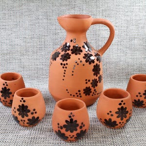 May include: Set of six terracotta pottery cups and a jug with black floral designs. The cups and jug are all decorated with black dots and flowers.