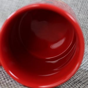 Peut inclure: Une tasse en c&eacute;ramique rouge avec une finition brillante. L'int&eacute;rieur de la tasse est rouge fonc&eacute; et l'anse est visible sur le c&ocirc;t&eacute; gauche. La tasse est pos&eacute;e sur un fond gris textur&eacute;.