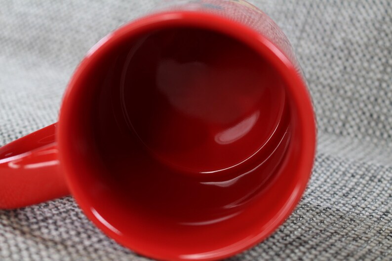 Peut inclure: Une tasse en c&eacute;ramique rouge avec une finition brillante. L'int&eacute;rieur de la tasse est rouge fonc&eacute; et l'anse est visible sur le c&ocirc;t&eacute; gauche. La tasse est pos&eacute;e sur un fond gris textur&eacute;.