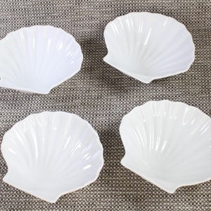 May include: Four white, shell-shaped dishes on a grey textured surface. Each dish has a ribbed design and a glossy finish. These decorative dishes are suitable for serving small portions of food or as decorative accents.