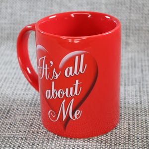 Peut inclure: Une tasse en c&eacute;ramique rouge vif avec une anse incurv&eacute;e. La tasse pr&eacute;sente un motif de c&oelig;ur blanc avec le texte "It's all about Me" dans une police &eacute;l&eacute;gante. La tasse est cylindrique et repose sur une surface textur&eacute;e.