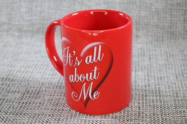 Peut inclure: Une tasse en c&eacute;ramique rouge vif avec une anse incurv&eacute;e. La tasse pr&eacute;sente un motif de c&oelig;ur blanc avec le texte "It's all about Me" dans une police &eacute;l&eacute;gante. La tasse est cylindrique et repose sur une surface textur&eacute;e.