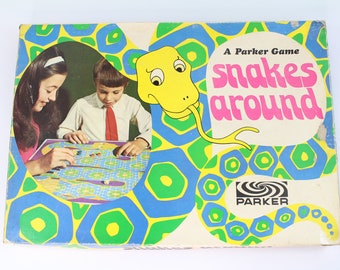 May include: A vintage board game box with a colourful design featuring a yellow snake with a blue and green background. The box reads "A Parker Game Snakes Around" with the Parker logo.
