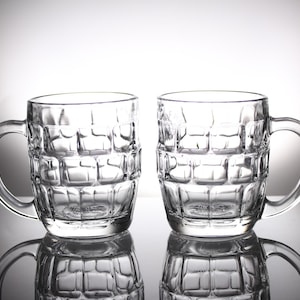 Op de afbeelding: Twee heldere glazen bierpullen met een getextureerd, vierkant patroon. Elke pul heeft een grote, gebogen handgreep. De pullen worden weerspiegeld in het glanzende oppervlak eronder, waardoor een symmetrisch beeld ontstaat. De achtergrond is een verloop van wit naar grijs.