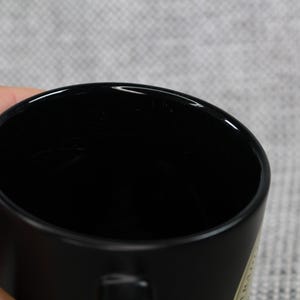 May include: A black ceramic mug with a glossy finish. The mug is held by a hand, with the interior of the mug visible. The background is a light gray fabric.