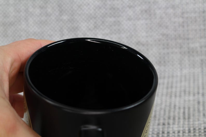 May include: A black ceramic mug with a glossy finish. The mug is held by a hand, with the interior of the mug visible. The background is a light gray fabric.