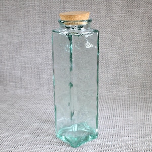 May include: A tall, square-shaped clear glass bottle with a textured, slightly wavy surface. The bottle features a thick, light green tint and is topped with a cylindrical, natural-coloured cork stopper that fits securely into the bottle opening.