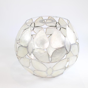 Può includere: Un paralume sferico con una struttura in metallo argentato e intarsi bianchi a forma di petalo. Il paralume ha una forma bulbosa ed è progettato per diffondere la luce. Gli intarsi sono disposti in un motivo sovrapposto.