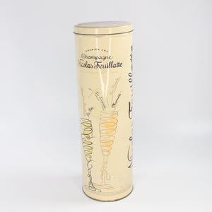 Könnte beinhalten: Ein hoher, zylindrischer Champagnerflaschenbehälter mit cremefarbenem Hintergrund. Der Behälter trägt den Text "Champagne Nicolas Feuillatte" und eine Zeichnung eines Sektglases. Das Design umfasst schwarze und goldene Akzente.