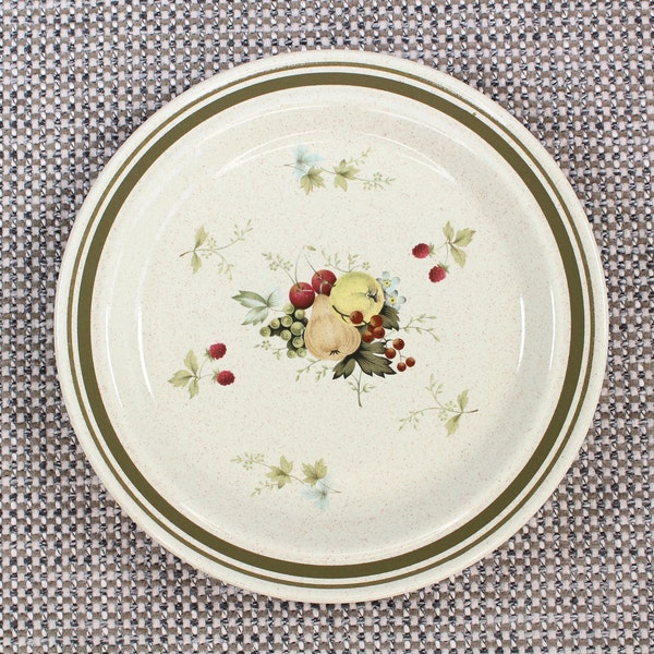 May include: A white ceramic plate with a green rim and a floral design featuring a pear, grapes, and other fruits. The plate has a speckled texture.