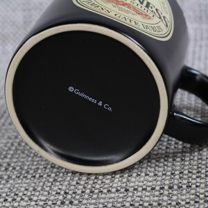 May include: A black ceramic Guinness mug with a white rim and handle. The mug features the Guinness logo and text on the side. The bottom of the mug has the text "&copy; Guinness & Co."
