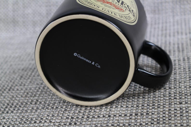 May include: A black ceramic Guinness mug with a white rim and handle. The mug features the Guinness logo and text on the side. The bottom of the mug has the text "&copy; Guinness & Co."