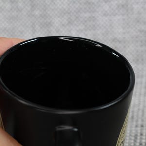May include: A black ceramic mug with a glossy finish. The mug is held by a hand, with the interior of the mug appearing dark. The mug has a simple, classic design, suitable for coffee or tea.