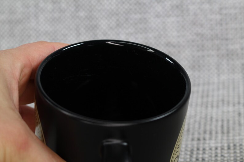 May include: A black ceramic mug with a glossy finish. The mug is held by a hand, with the interior of the mug appearing dark. The mug has a simple, classic design, suitable for coffee or tea.