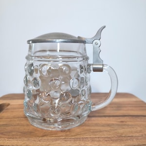 以下が含まれることがあります： ディンプル模様の透明なガラス製ビールジョッキ。金属製のヒンジ付き蓋とハンドルが付いています。ジョッキは木製のまな板の上に置かれています。蓋とハンドルは銀色の金属製です。