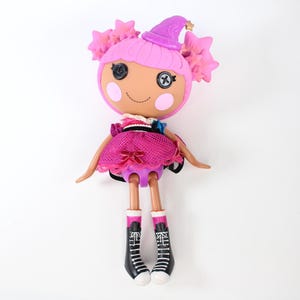 May include: A pink and purple doll with a witch hat and star details. The doll has a round face with button eyes and a stitched smile. It wears a pink dress and black and white boots. The doll's arms and legs are poseable.