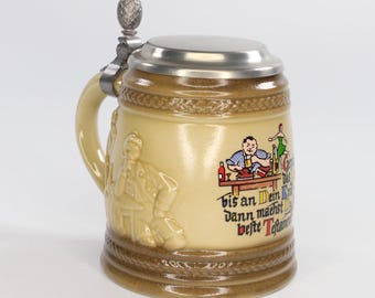 Op de afbeelding: Een bruin keramische bierpul met een metalen deksel en handvat. De pul heeft een reliëf van een man en een kleurrijke afbeelding met tekst. De basis heeft een decoratieve band.