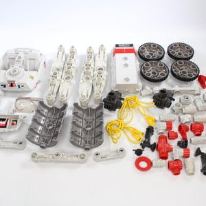 May include: A selection of disassembled toy robot components in white, grey, red, and black. The parts include wheels, connectors, and body panels, indicating a construction or building toy. Various sizes and shapes are visible.