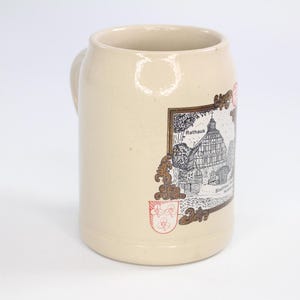 May include: A beige ceramic beer stein with a handle. The stein features a detailed illustration of a building labeled "Rathaus" within an ornate frame. A red emblem is below the frame. The stein is a traditional drinking vessel.