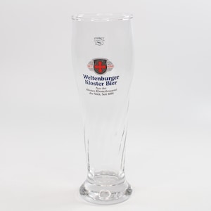 Könnte beinhalten: Ein hohes, klares Bierglas mit ausgestelltem Rand und rundem Boden. Das Glas zeigt die Aufschrift "Weltenburger Kloster Bier" in Blau und Rot, ein Wappen und den Text "Seit 1050".