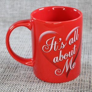 Peut inclure: Une tasse en c&eacute;ramique rouge avec une anse incurv&eacute;e. La tasse porte l'inscription "It's all about Me" en lettres blanches &agrave; l'int&eacute;rieur d'un c&oelig;ur. La tasse est de couleur rouge uni.