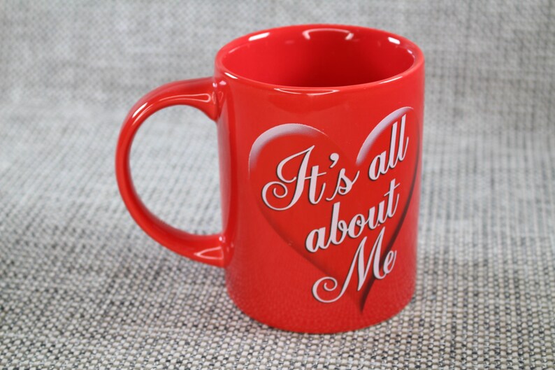 Peut inclure: Une tasse en c&eacute;ramique rouge avec une anse incurv&eacute;e. La tasse porte l'inscription "It's all about Me" en lettres blanches &agrave; l'int&eacute;rieur d'un c&oelig;ur. La tasse est de couleur rouge uni.