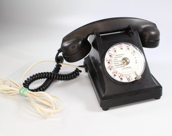 May include: A black rotary dial phone with a white dial face and a black coiled cord. The dial face has red numbers and letters and text in French.
