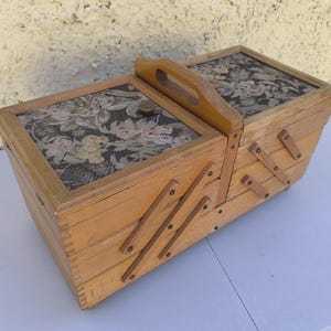 May include: A wooden sewing box with a cantilevered accordion design and a central handle. The two top lids feature a dark fabric inlay with a floral pattern in shades of pink, green, and cream. The box has multiple tiers and wooden joints.