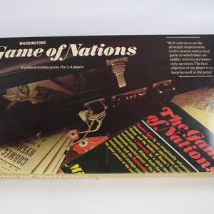 Könnte beinhalten: Vintage Brettspiel "Game of Nations" von Waddingtons. Die Box hat ein schwarzes Aktentaschendesign mit goldenen Details und dem Spieltitel in Weiß. Enthält Text über das Ziel des Spiels und ein politisches Strategiethema.