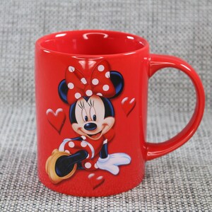 Peut inclure: Une tasse en c&eacute;ramique rouge repr&eacute;sentant Minnie Mouse. La tasse a une grande anse et un motif de Minnie Mouse portant un n&oelig;ud et une robe &agrave; pois. Des pois blancs sont sur le n&oelig;ud et la robe. Des c&oelig;urs sont &eacute;galement sur la tasse.