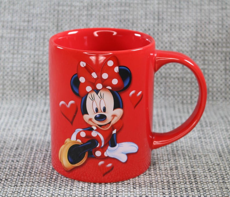 Peut inclure: Une tasse en c&eacute;ramique rouge repr&eacute;sentant Minnie Mouse. La tasse a une grande anse et un motif de Minnie Mouse portant un n&oelig;ud et une robe &agrave; pois. Des pois blancs sont sur le n&oelig;ud et la robe. Des c&oelig;urs sont &eacute;galement sur la tasse.