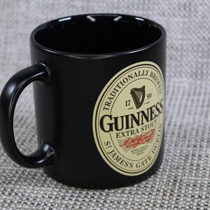 May include: Black ceramic mug with a curved handle. The mug features the Guinness logo, a cream-colored oval with the words "Guinness Extra Stout" and "St. James's Gate Dublin" printed on it. The mug is a classic style.