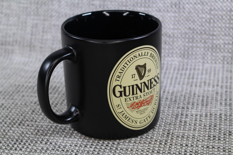 May include: Black ceramic mug with a curved handle. The mug features the Guinness logo, a cream-colored oval with the words "Guinness Extra Stout" and "St. James's Gate Dublin" printed on it. The mug is a classic style.