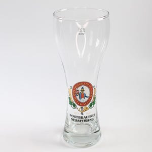 Könnte beinhalten: Ein klares, hohes Bierglas mit ausgestelltem Rand und rundem Boden. Das Glas hat ein farbenfrohes rundes Emblem mit einer Figur in traditioneller Kleidung, dem Text "POSTBRAUEREI NESSELWANG" und "SEIT 1650". Das Glas hat ein Fassungsvermögen von 0,2 l.