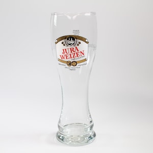 Puede incluir: Una copa de cerveza de vidrio transparente con forma acampanada y una etiqueta decorativa. La etiqueta presenta el texto "JURA WEIZEN" en rojo y blanco, con un borde dorado y marrón. La copa tiene una capacidad de 120 ml.