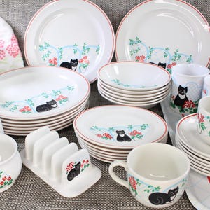 May include: A set of white ceramic plates and mugs featuring black cat illustrations and red floral accents. The plates have a blue and white pattern with a cat sitting on a branch. The mugs feature a black cat with a white chest and a red flower behind it. The set includes a salt and pepper shaker, a bread knife holder, and a teacup and saucer.
