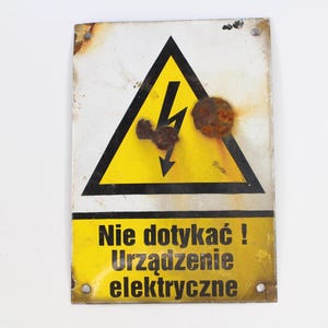 May include: A rectangular metal warning sign featuring a yellow triangle with a black lightning bolt symbol. The sign has black text that reads "Nie dotykać! Urządzenie elektryczne" and shows signs of rust and wear.