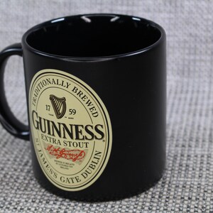 May include: Black ceramic mug with a curved handle. The mug features a Guinness Extra Stout label with a harp, the words "Traditionally Brewed", and "James's Gate Dublin".