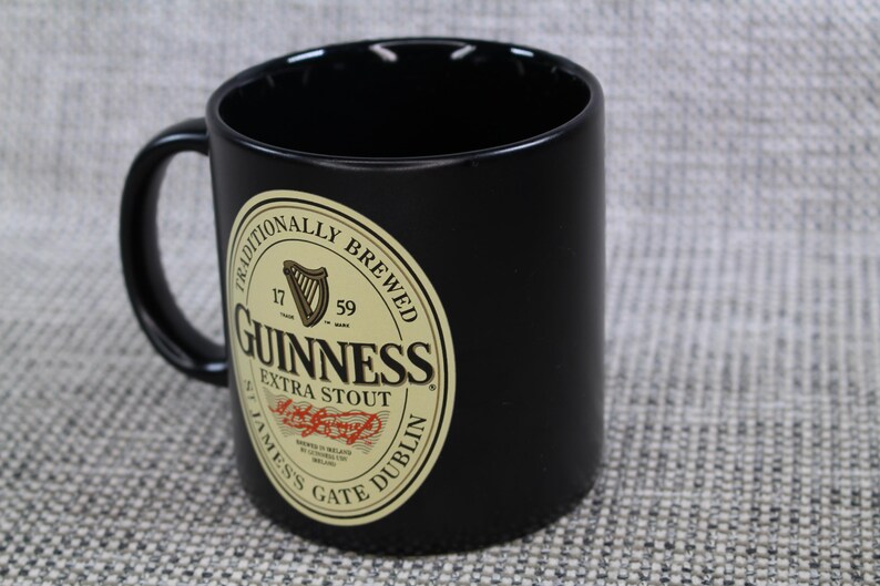 May include: Black ceramic mug with a curved handle. The mug features a Guinness Extra Stout label with a harp, the words "Traditionally Brewed", and "James's Gate Dublin".