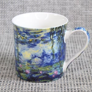 Pode incluir: Uma caneca de cerâmica com uma pintura de nenúfares em tons de azul, verde e amarelo. A alça e a borda são brancas, e o interior também é branco. A caneca tem um tamanho padrão para bebidas quentes.