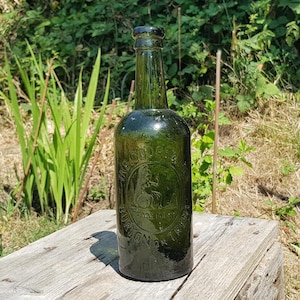 May include: A vintage green glass bottle with embossed lettering and a circular emblem. The bottle is cylindrical with a long neck and sits on a weathered wooden surface. The bottle is outdoors in natural light.