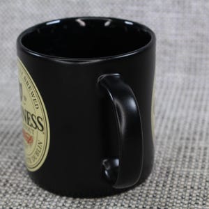 May include: Black ceramic mug with a curved handle. The mug features a circular label with the word "Guinness" and other text. The mug is designed for beverages and is suitable for home or office use.