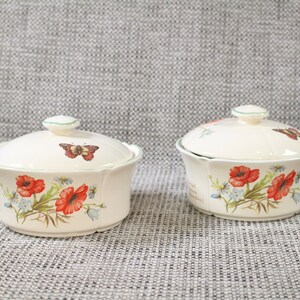 May include: Two white ceramic bowls with lids, featuring a floral design with red poppies, green leaves, and butterflies. The bowls have a scalloped edge and a green trim.