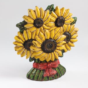 May include: A decorative bouquet of sunflowers with yellow petals and dark brown centers. The sunflowers are tied with a red bow and green leaves. The bouquet is a decorative item.