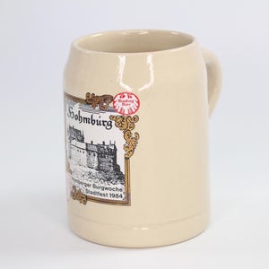 May include: A beige ceramic beer stein with a handle. The stein features a decorative label with the text "Sohmburg" and a castle illustration. The label also includes the text "Binding Bier" and "Nürnberger Burgwoche Stadtfest 1984".