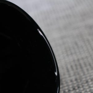 May include: Close-up of a glossy black ceramic bowl. The bowl's interior is dark, reflecting light. The rim of the bowl is visible, with a smooth, curved edge. The background is a textured, light gray surface.