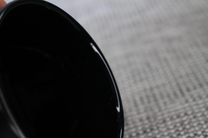 May include: Close-up of a glossy black ceramic bowl. The bowl's interior is dark, reflecting light. The rim of the bowl is visible, with a smooth, curved edge. The background is a textured, light gray surface.