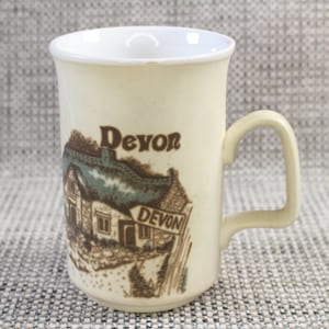 May include: A white ceramic mug with a brown and green illustration of a cottage with a thatched roof and a sign that reads "Devon". The mug has the word "Devon" written in brown lettering on the front.